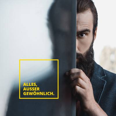 Porträt eines bärtigen Mannes, der hinter einer dunklen Fläche hervorschaut, mit dem gelben Casa Nova Slogan ‚Alles, außer gewöhnlich.‘ im Vordergrund