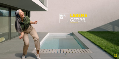 Eine glückliche, ältere Frau mit Kopfhörern tanzt auf der modernen Steinterrasse eines Casa Nova Architektenhauses neben einem Pool mit dem Slogan ‚Lebensgefühl‘