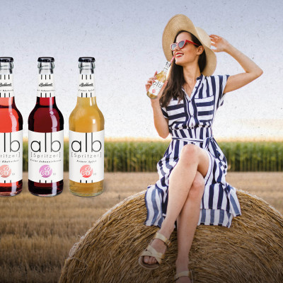 Werbemotiv von Burkhardt Fruchtsäfte: Eine Frau mit Sonnenhut und gestreiftem Kleid sitzt auf einem Strohballen und trinkt eine alb Schorle, daneben die Sorten Rhabarber, Johannisbeere und Apfel