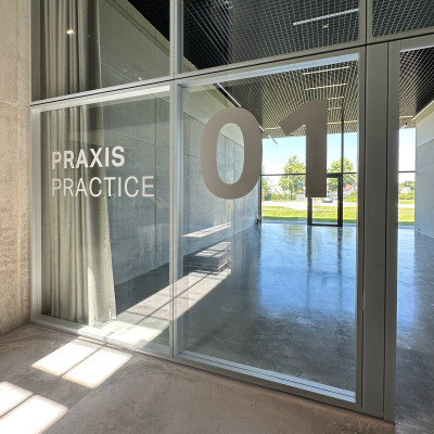 Blick durch eine großflächige Glaswand auf den Praxisraum mit Signaletik ‚PRAXIS / PRACTICE 01‘ im Geberit Campus, mit spiegelndem Industrieboden und Blick ins Grüne