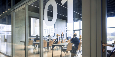 Blick durch eine Glaswand in den modernen Seminarraum ‚THEORIE 01 / THEORY 01‘ im Geberit Campus, ausgestattet mit einer großen LED-Wand, langen Tischen und Sitzreihen