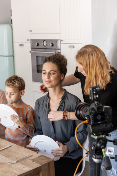 Behind-the-Scenes-Aufnahme eines Werbedrehs für tellofix: Eine Stylistin richtet die Haare einer Frau, während diese und ein Kind an einem Küchentisch sitzen und leere Teller halten; im Vordergrund ist eine Filmkamera auf einem Stativ zu sehen