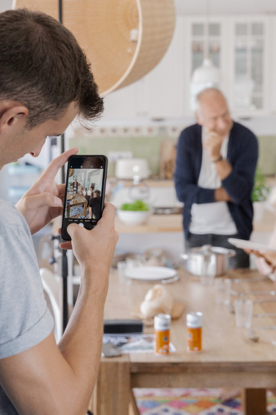Blick über die Schulter eines Kameramanns, der mit einem Smartphone eine Kochszene für tellofix filmt; im unscharfen Hintergrund steht ein älterer Mann in einer hellen Küche am Esstisch